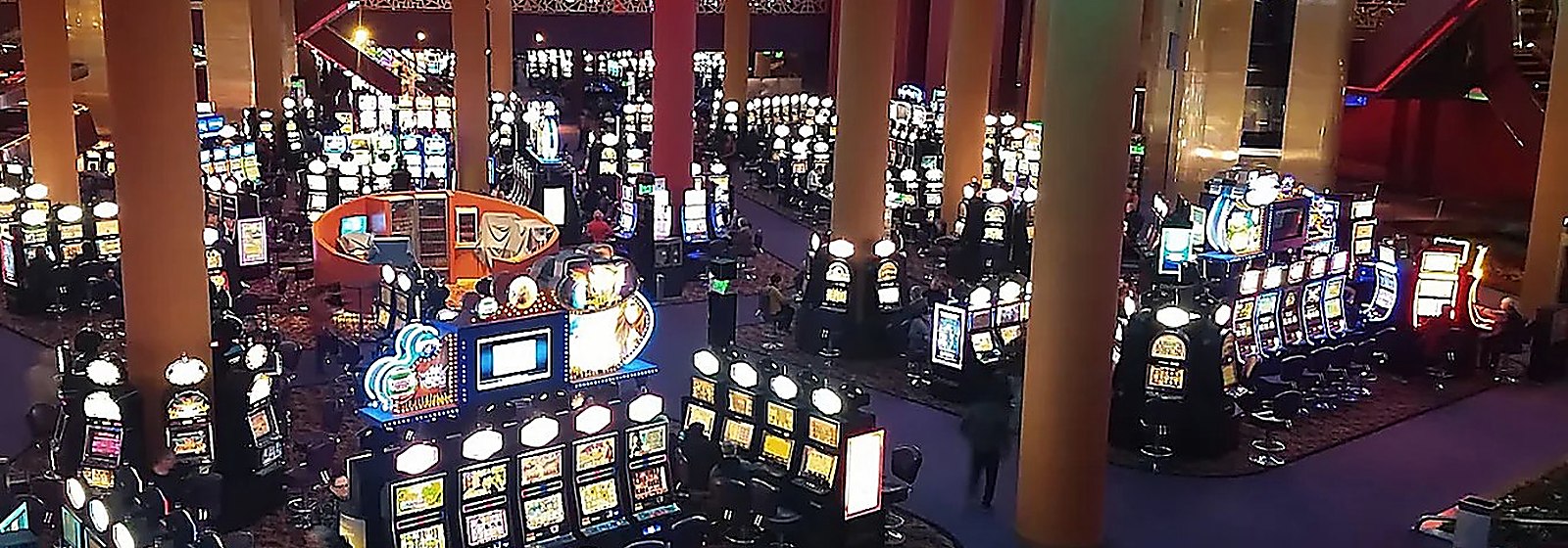 Salón de juego amplio visto desde un entrepiso, con hileras de máquinas iluminadas, columnas anaranjadas de doble altura y luces cenitales.